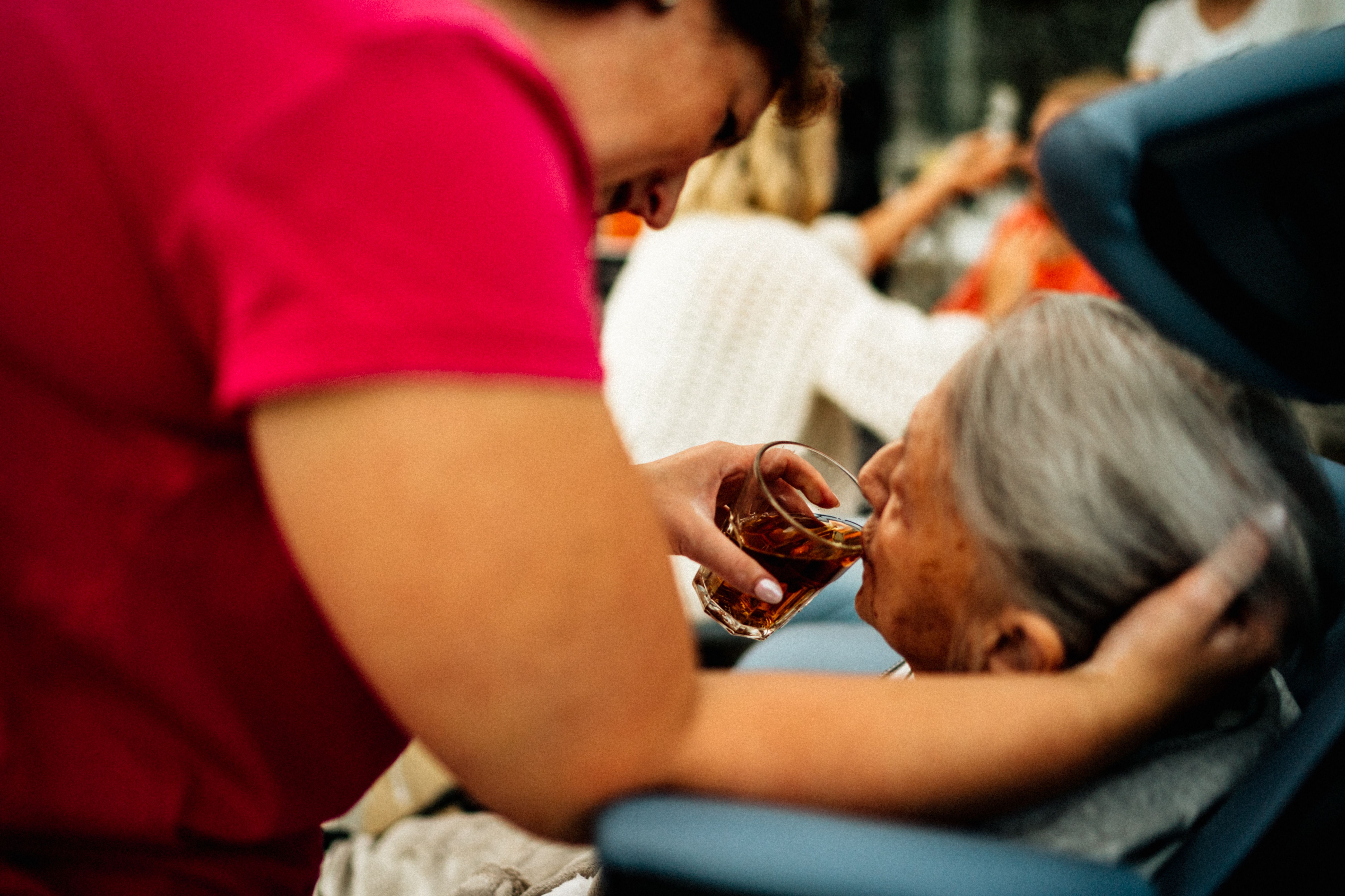 A woman wearing red scrubs holds a glass for an elderly woman in a wheelchair, allowing her to drink.